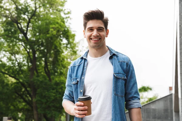 Bild eines männlichen jungen Mannes, der Kaffee zum Mitnehmen trinkt und einen Laptop in der Hand hält, während er durch den grünen Park geht — Stockfoto