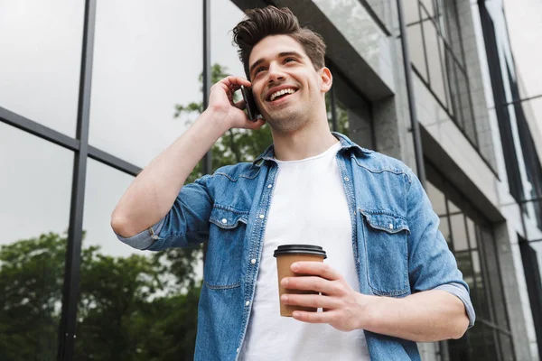 Imagen de un joven moreno bebiendo café para llevar y hablando por celular mientras está de pie sobre el edificio — Foto de Stock