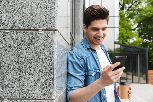 Gelukkig jong geweldig man zakenman poseren buitenshuis buiten wandelen chatten door mobiele telefoon drinken koffie. — Stockfoto