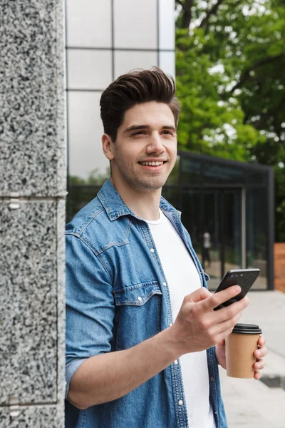 Happy young amazing man businessman posing outdoors outside walking chatting by mobile phone drinking coffee.