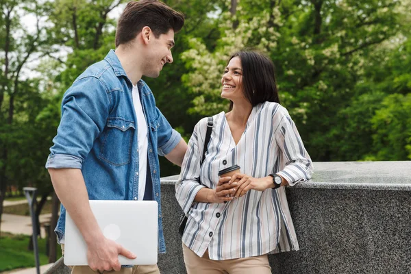 Image de couple attrayant avec tasse en papier et ordinateur portable parlant tout en se tenant debout sur les escaliers à l'extérieur — Photo