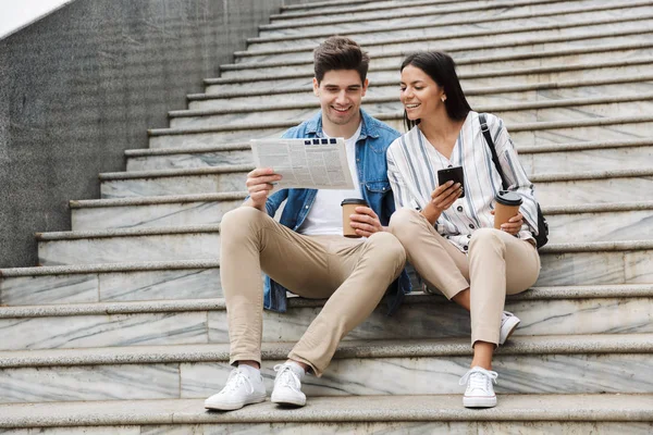 Happy young amazing loving couple business people colleagues outdoors outside on steps reading newspaper drinking coffee.