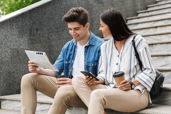 Happy young amazing loving couple business people colleagues outdoors outside on steps reading newspaper drinking coffee.