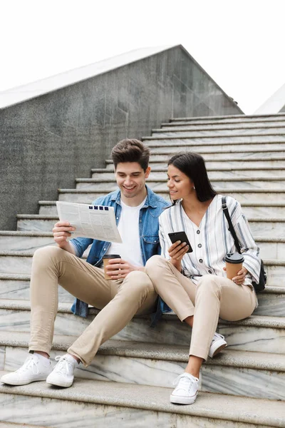 Happy young amazing loving couple business people colleagues outdoors outside on steps reading newspaper drinking coffee. — Stock Photo, Image