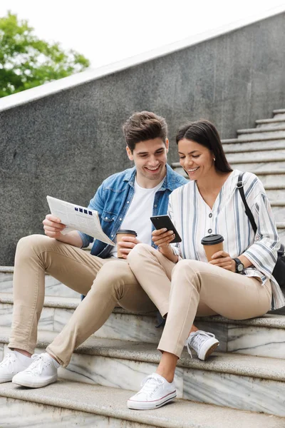 Feliz joven increíble pareja amorosa gente de negocios colegas al aire libre al aire libre en los pasos leyendo periódico beber café . — Foto de Stock