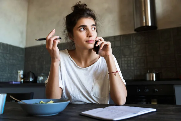 Fotó a komor barna nő írásban naplót, és beszél a mobiloddal, miközben reggeli konyhában — Stock Fotó