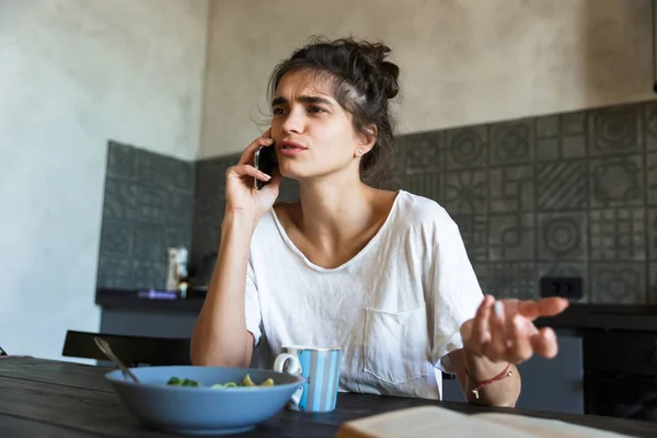 Photo of brooding brunette woman reading book and talking on cellphone while having breakfast in kitchen