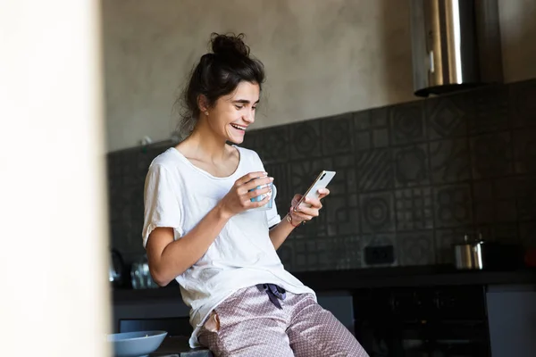 Fotó szép nevetve nő visel otthon ruhákat iszik kávét gépelés közben a mobiltelefon a konyhában — Stock Fotó