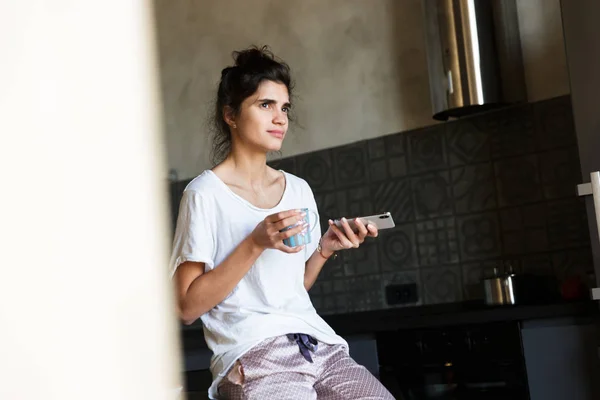 Photo of cute thinking woman wearing leisure clothes drinking coffee while holding cellphone in kitchen