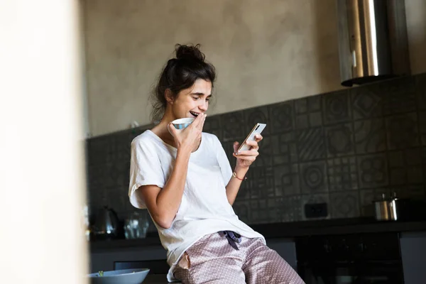 Fotó aranyos nevetés nő visel szabadidő ruhák kávét, miközben tartja cellphone a konyhában — Stock Fotó