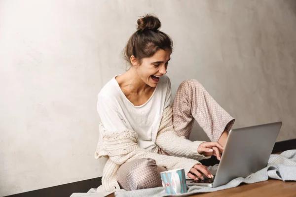 Foto de la feliz mujer riendo bebiendo café y utilizando el ordenador portátil mientras está sentado en el suelo en casa — Foto de Stock