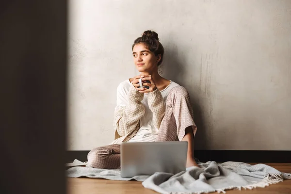 Foto de mujer de ensueño atractiva bebiendo café y usando el ordenador portátil mientras está sentado en el suelo en casa — Foto de Stock