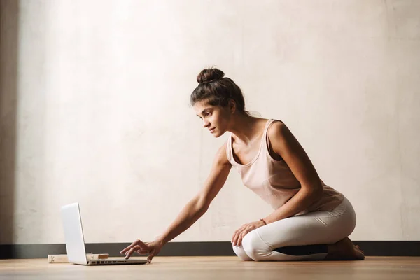Foto di focalizzata bella donna in abbigliamento sportivo digitando sul computer portatile mentre seduto sul pavimento — Foto Stock