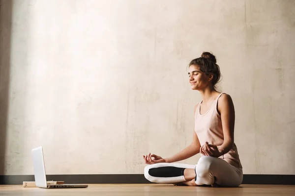 Foto av Joyful Fitness kvinna mediterar med Zen fingrar och leende medan du sitter på golvet med laptop hemma — Stockfoto