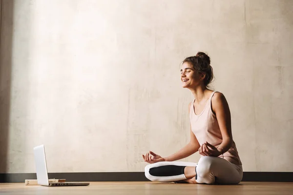 Foto av glada Fitness kvinna mediterar med Zen fingrar och skrattar medan du sitter på golvet med laptop hemma — Stockfoto