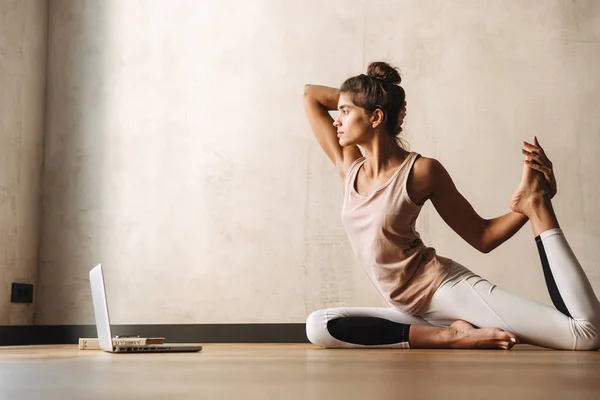 Foto av ganska koncentrerad kvinna gör yoga övningar med hjälp av laptop medan du sitter på golvet hemma — Stockfoto