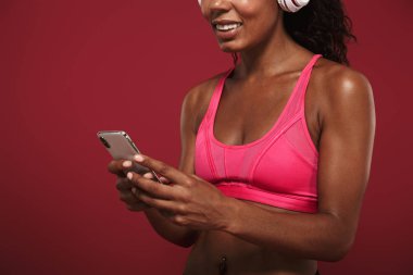 Gülümseyen memnun güçlü genç afrika fitness spor kadın kulaklık ile müzik dinleyerek kırmızı duvar arka plan üzerinde izole poz.