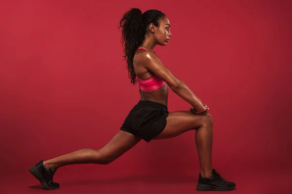 Концентрована сильна молода африканська спортивна жінка позує ізольовано на фоні червоної стіни робить спортивні вправи . — стокове фото