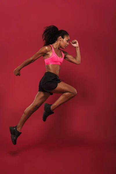 Corredor concentrado fuerte joven africano deportes mujer posando aislado sobre rojo pared fondo corriendo saltar . — Foto de Stock