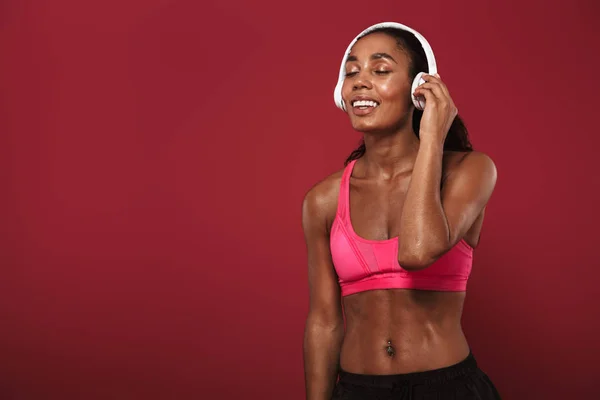 Lächelnd erfreut starke junge afrikanische Fitness-Sportlerin posiert isoliert über roter Wand Hintergrund Musik hören mit Kopfhörern. — Stockfoto