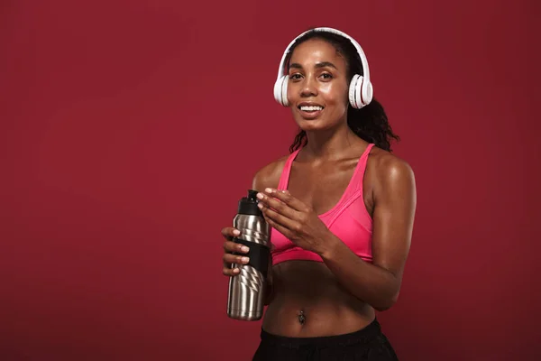 Mosolyogva tetszett erős fiatal afrikai fitness sport nő jelentő elszigetelt mint a vörös fal háttérben hallgat zenét fejhallgató. — Stock Fotó
