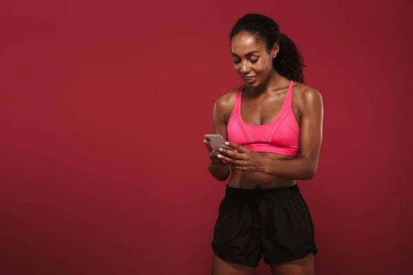 Erős fiatal afrikai fitnesz sport nő pózol elszigetelt vörös fal háttérben a mobiltelefon csevegés. — Stock Fotó