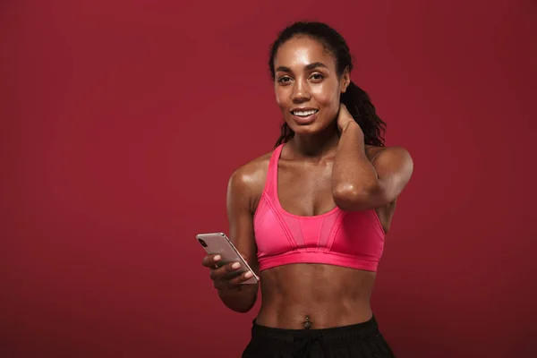Örömmel mosolygó pozitív erős fiatal afrikai fitnesz sport nő pózol elszigetelt vörös fal háttérben a mobiltelefon csevegés. — Stock Fotó