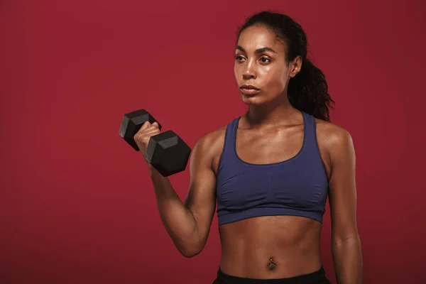 Sério forte jovem Africano fitness esportes mulher posando isolado sobre fundo da parede vermelha fazer exercício para os braços com halteres . — Fotografia de Stock