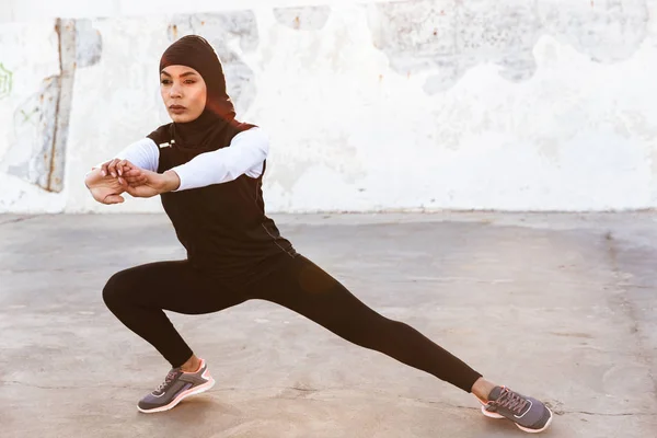 Μουσουλμανική αθλητική γυναίκα γυμναστήριο ντυμένη σε χιτζάμπ και σκούρα ρούχα που θέτουν κάνει άθλημα τέντωμα ασκήσεις σε εξωτερικούς χώρους στο δρόμο. — Φωτογραφία Αρχείου
