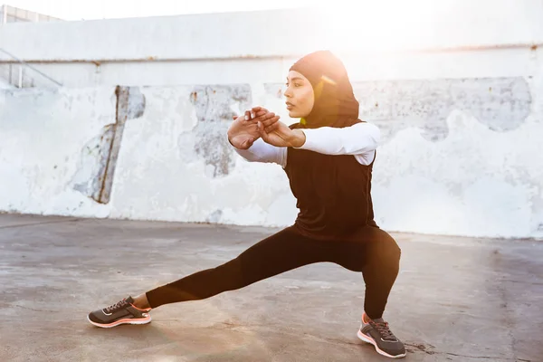 Ισχυρή μουσουλμανική γυμναστήριο γυναίκα ντυμένη σε χιτζάμπ και σκούρα ρούχα που θέτουν κάνει άθλημα τέντωμα ασκήσεις σε εξωτερικούς χώρους στο δρόμο. — Φωτογραφία Αρχείου