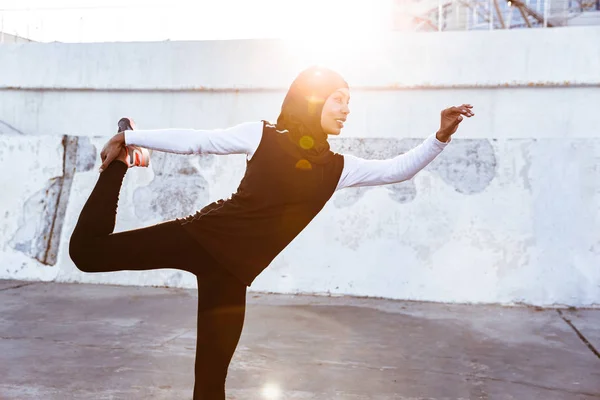 Ισχυρή μουσουλμανική γυμναστήριο γυναίκα ντυμένη σε χιτζάμπ και σκούρα ρούχα που θέτουν κάνει άθλημα τέντωμα ασκήσεις σε εξωτερικούς χώρους στο δρόμο. — Φωτογραφία Αρχείου