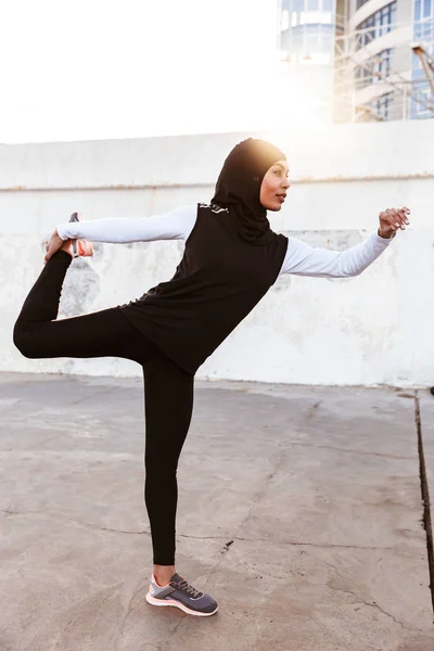 Skoncentrowany muzułmański Sport fitness kobieta ubrana w hidżab i ciemne ubrania stwarzające zrobić sportowe ćwiczenia rozciągające na zewnątrz na ulicy. — Zdjęcie stockowe
