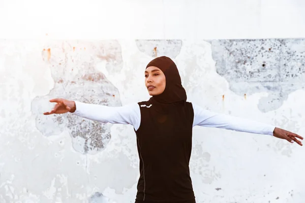 Skoncentrowany muzułmańskich sportu fitness kobieta ubrana w hidżab i ciemne ubrania stwarzające sprawiają, że ćwiczenia rozciągające Sport na zewnątrz na ulicy zrobić ćwiczenia jogi. — Zdjęcie stockowe