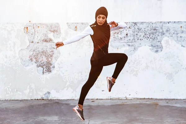 Koncentrerad Muslim sport Fitness kvinna klädd i hijab och mörka kläder poserar göra sport stretching övningar utomhus på gatan som kör. — Stockfoto