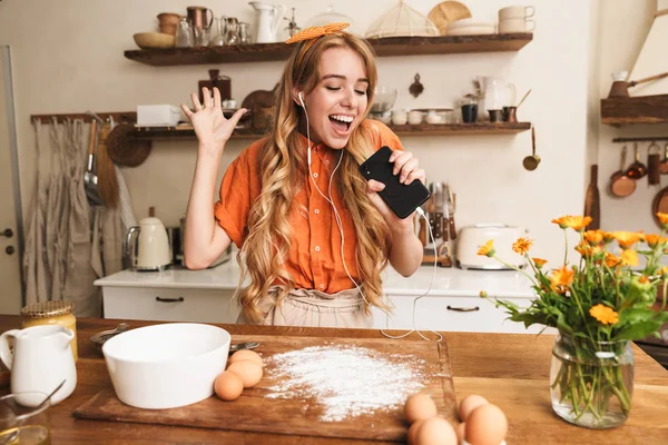 Örömmel optimista fiatal szőke lány szakács főzés a konyhában hallgat zenét fülhallgató segítségével mobiltelefon énekel. — Stock Fotó