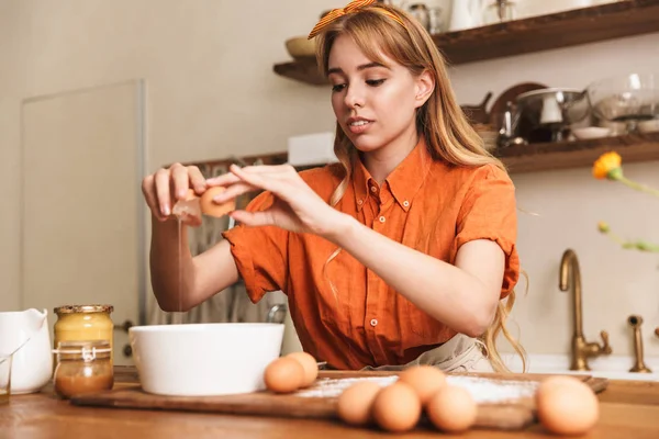 Konsantre genç sarışın kız şef yumurta ile mutfakta yemek. — Stok fotoğraf