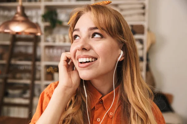 Felice ottimista giovane ragazza bionda in camicia arancione in cucina ascoltando musica con gli auricolari . — Foto Stock