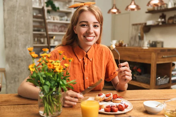 Lykkelig ung blond jente på kjøkkenet spiser søt konditorkaker spiser frokost og drikker juice . – stockfoto