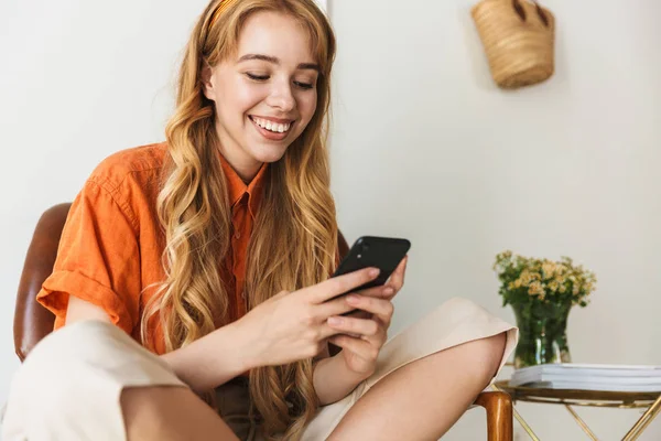 Glimlachend jong blond meisje thuis binnenshuis met behulp van mobiele telefoon zittend op stoel. — Stockfoto