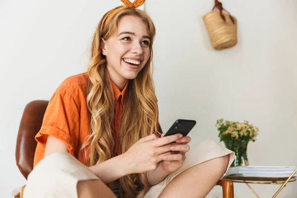 Usmívá se Mladá blondýnka doma a používá mobilní telefon sedící na židli. — Stock fotografie