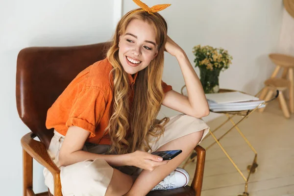 Усміхнена молода блондинка вдома використовує мобільний телефон, сидячи на стільці . — стокове фото