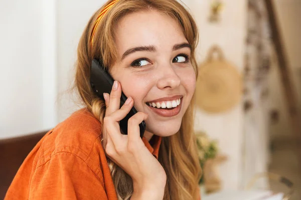 Glimlachend jong blond meisje thuis binnenshuis praten door mobiele telefoon zittend op stoel. — Stockfoto