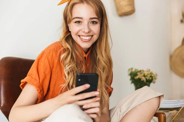 Усміхнена молода блондинка вдома використовує мобільний телефон, сидячи на стільці . — стокове фото