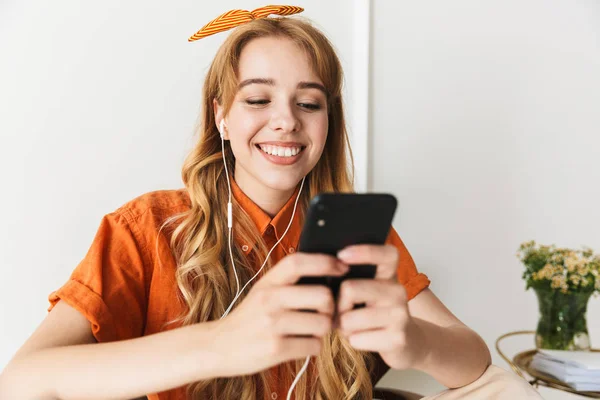 Glimlachend jong blond meisje thuis binnenshuis luisteren muziek met koptelefoon zittend op stoel. — Stockfoto