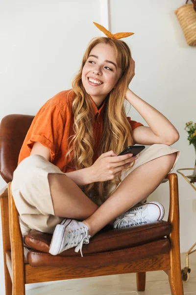 Hymyilevä nuori blondi tyttö kotona sisällä matkapuhelimella istuu tuolilla . — kuvapankkivalokuva
