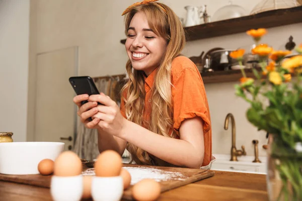 Mosolygó fiatal szőke lány szakács főzés a konyhában a mobiltelefon segítségével. — Stock Fotó