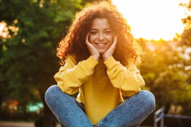 Mutlu mutlu sevimli genç öğrenci kıvırcık kız güzel güneş ışığı ile doğa parkı açık bankta oturan.