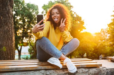Mutlu komik iyimser sevimli genç öğrenci kıvırcık kız cep telefonu kullanarak güzel güneş ışığı ile doğa parkı açık bankta oturan.