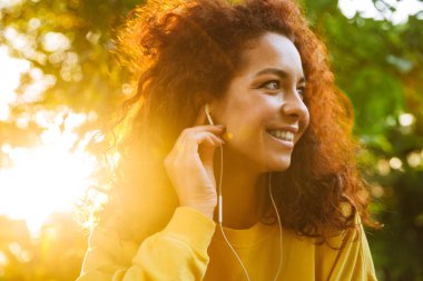 Mutlu iyimser sevimli genç öğrenci kıvırcık kız kulaklık ile müzik dinleyen güzel güneş ışığı ile doğa parkı açık tezgah üzerinde oturan.