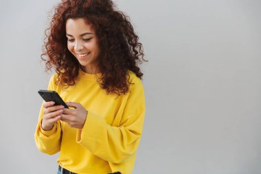 Mutlu gülümseyen neşeli güzel kıvırcık kız cep telefonu kullanarak gri arka plan üzerinde izole.